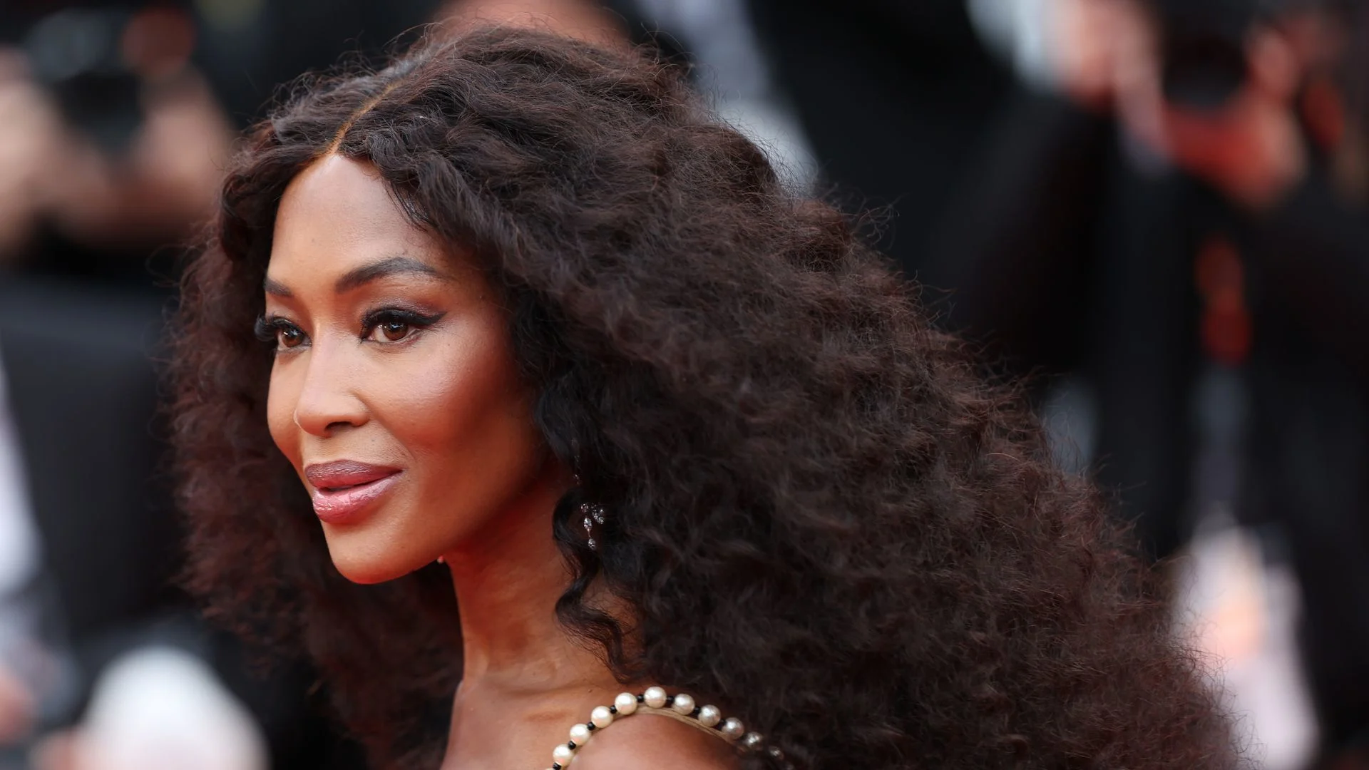Naomi Campbell smiling glamorously on the red carpet, wearing an elegant designer gown.
