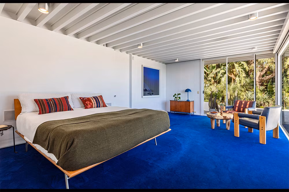 Primary bedroom in Brad Pitt’s former Steel House with cobalt-blue carpeting, large windows, and serene hillside views.