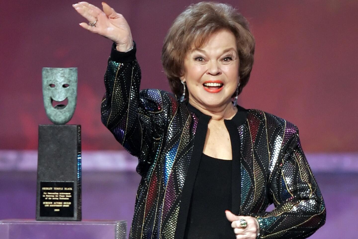 Shirley Temple as an adult waving to the crowd on stage at an awards show.