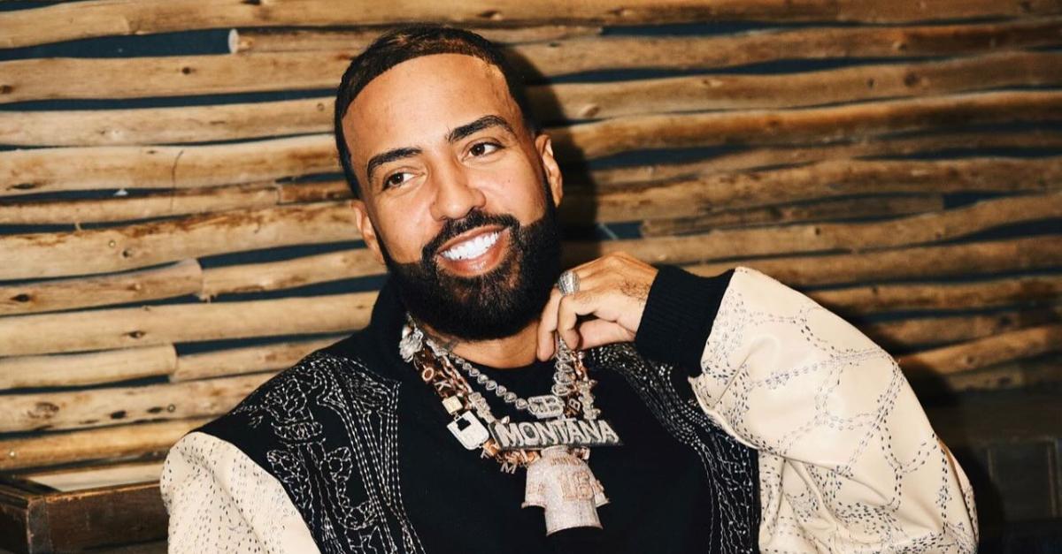 French Montana smiling and holding his gold chain during a public appearance.