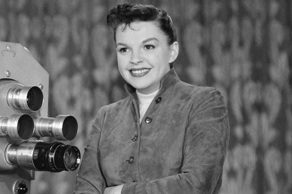 Judy Garland in black and white standing beside a vintage film camera, wearing a classic Hollywood outfit.
