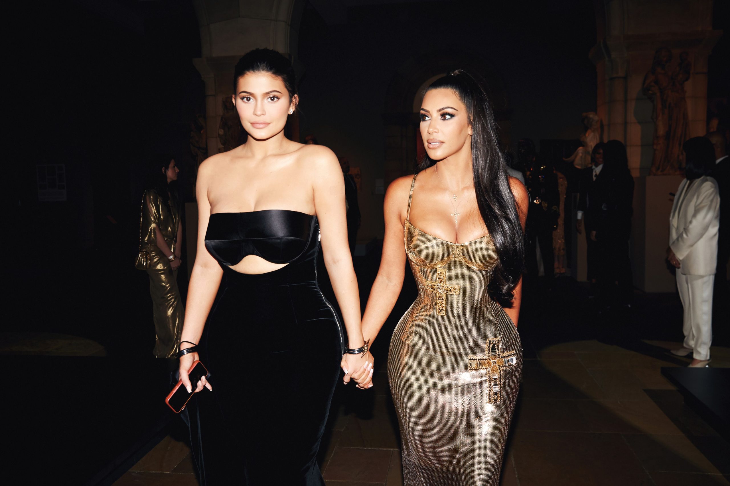 Kim Kardashian and Kylie Jenner dressed in glamorous outfits, holding hands and looking at the camera.