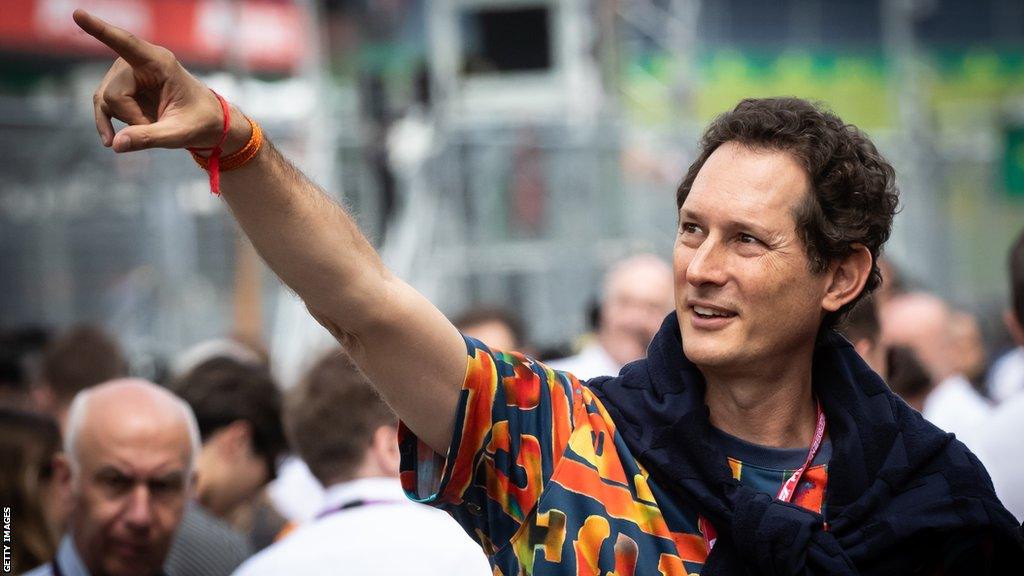 John Elkann pointing and waving to fans at a Formula 1 event, smiling and engaging with the crowd.