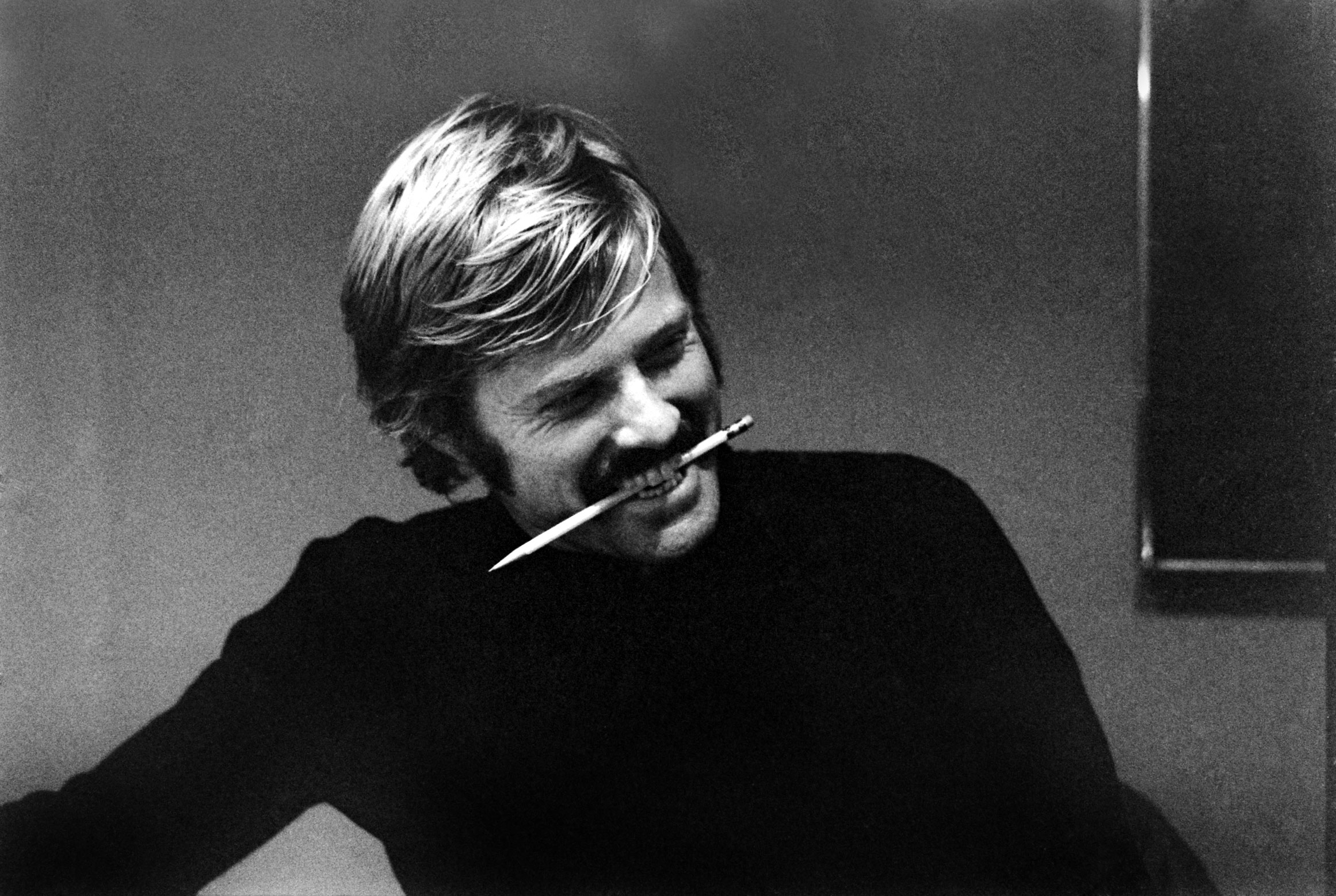 Young Robert Redford in black and white, smiling with a pencil in his mouth during a photo shoot.