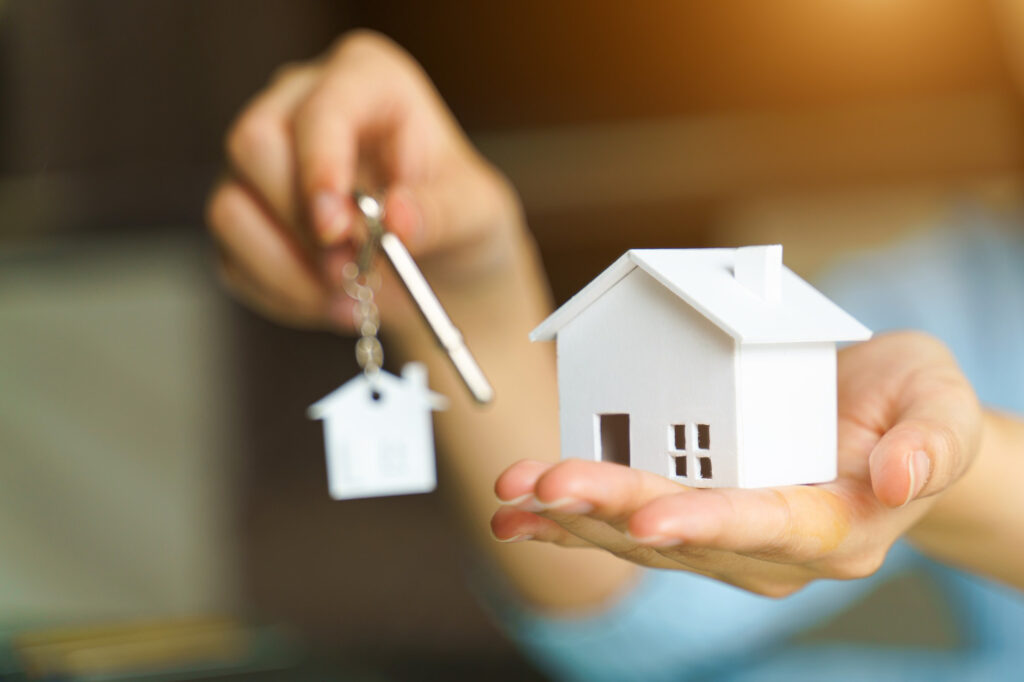 A hand holding a small wooden house along with keys, symbolizing homeownership and receiving a new mortgage.