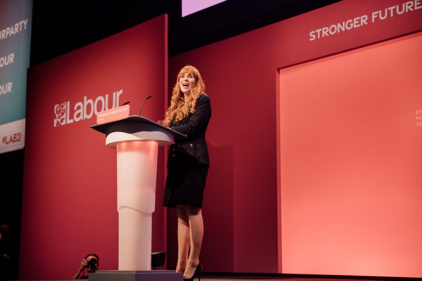 Angela Rayner delivering a passionate speech at the Labour Party Conference, standing at the podium with the Labour rose emblem behind her.