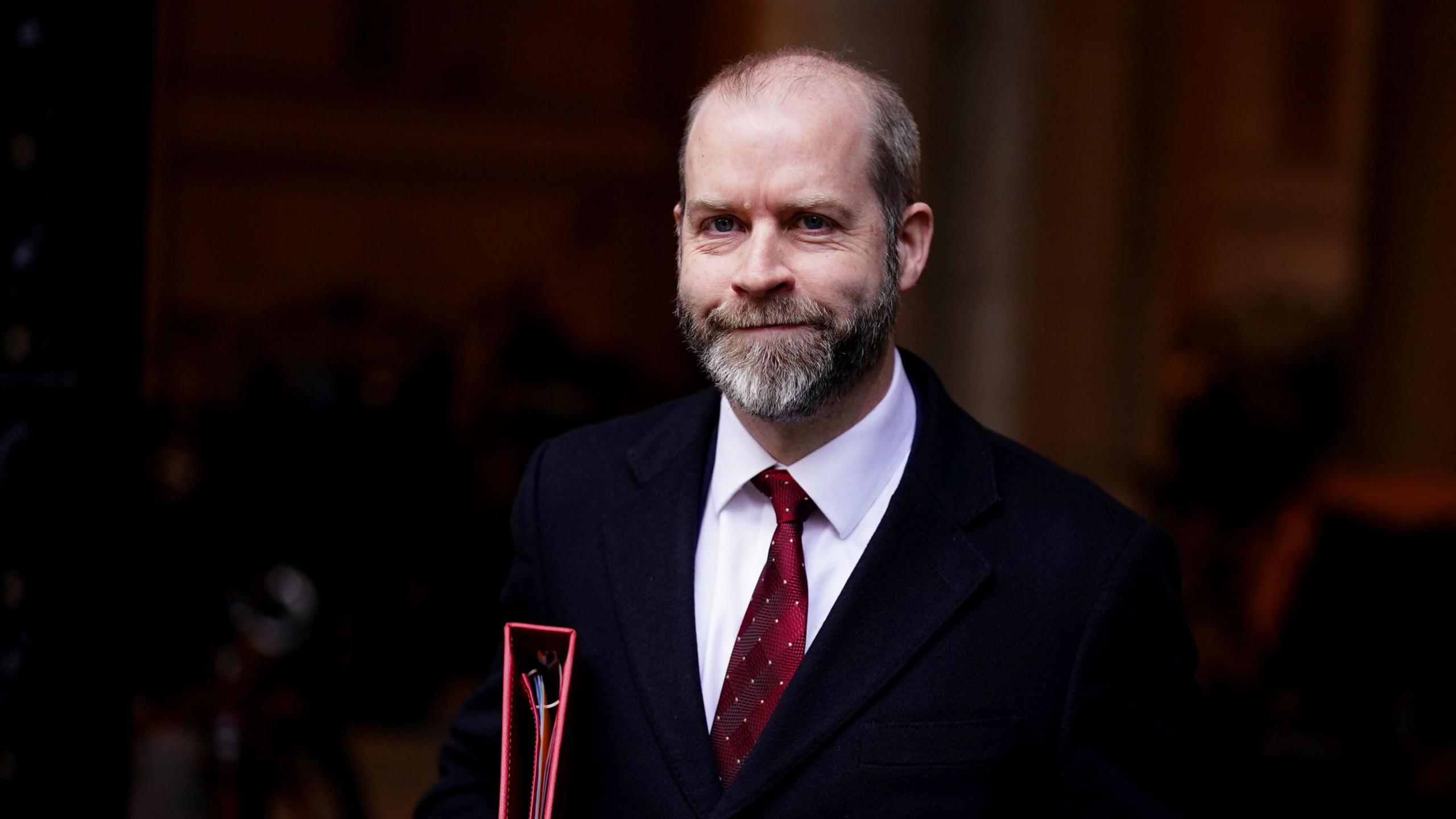 Jonathan Reynolds smiling at the camera, holding official documents under his arm in a professional setting.