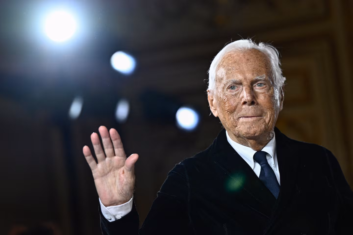 Giorgio Armani wearing a tailored suit, waving goodbye with a warm smile.