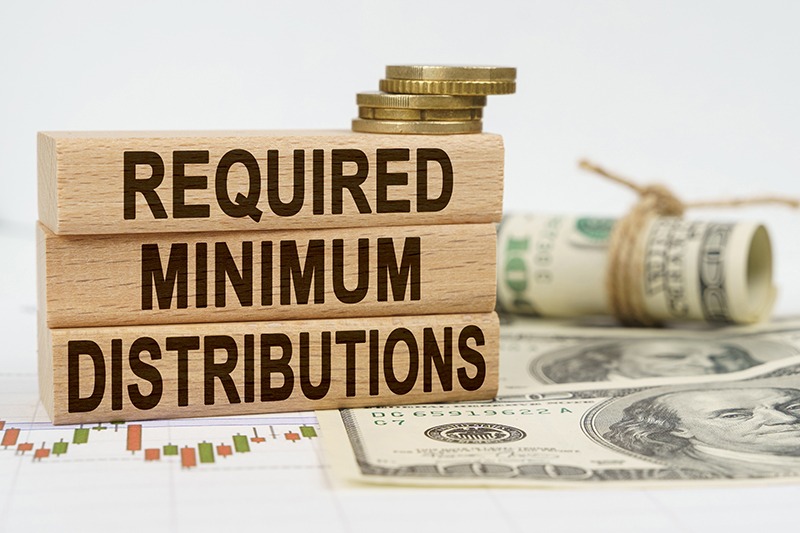 Wooden blocks spelling out “Required Minimum Distributions” with cash stacked behind them, symbolizing retirement account withdrawals and financial planning.