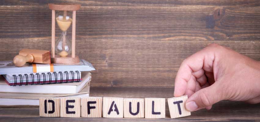 A hand arranging wooden letters to spell “DEFAULT” with a sand timer in the background, symbolizing time running out on loan repayment.