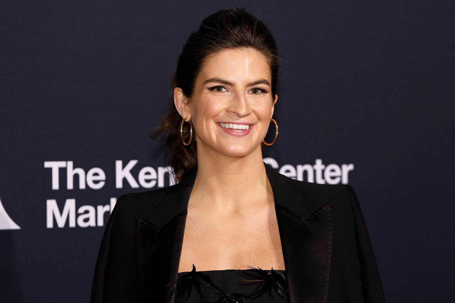 Kaitlan Collins attends a red carpet event, smiling in a black outfit with gold hoop earrings.