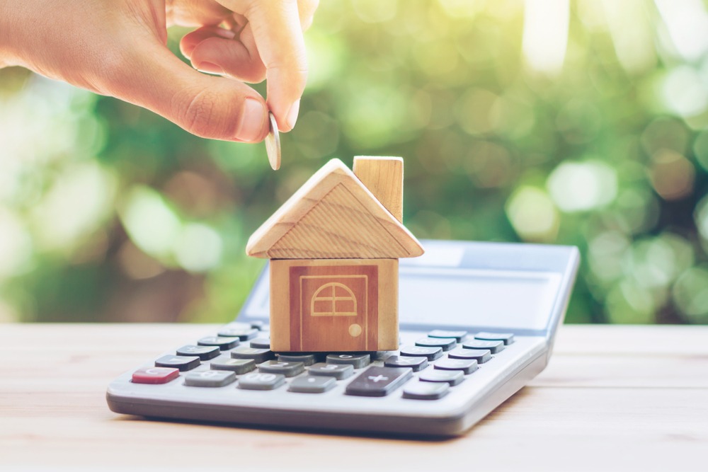 Person inserting money into a small wooden house-shaped money box placed on a calculator, symbolizing saving for a mortgage.