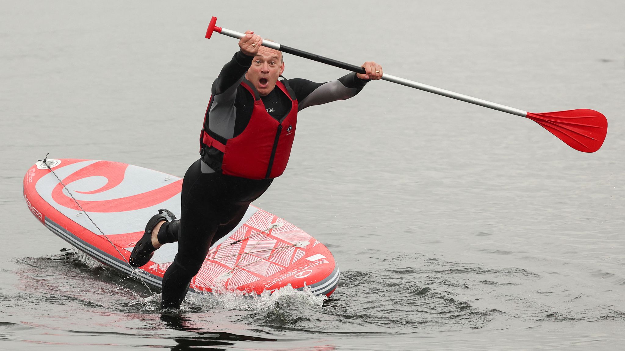 Ed Davey falling off a wave board during a public relations stunt, mid-action with water splashing around him.
