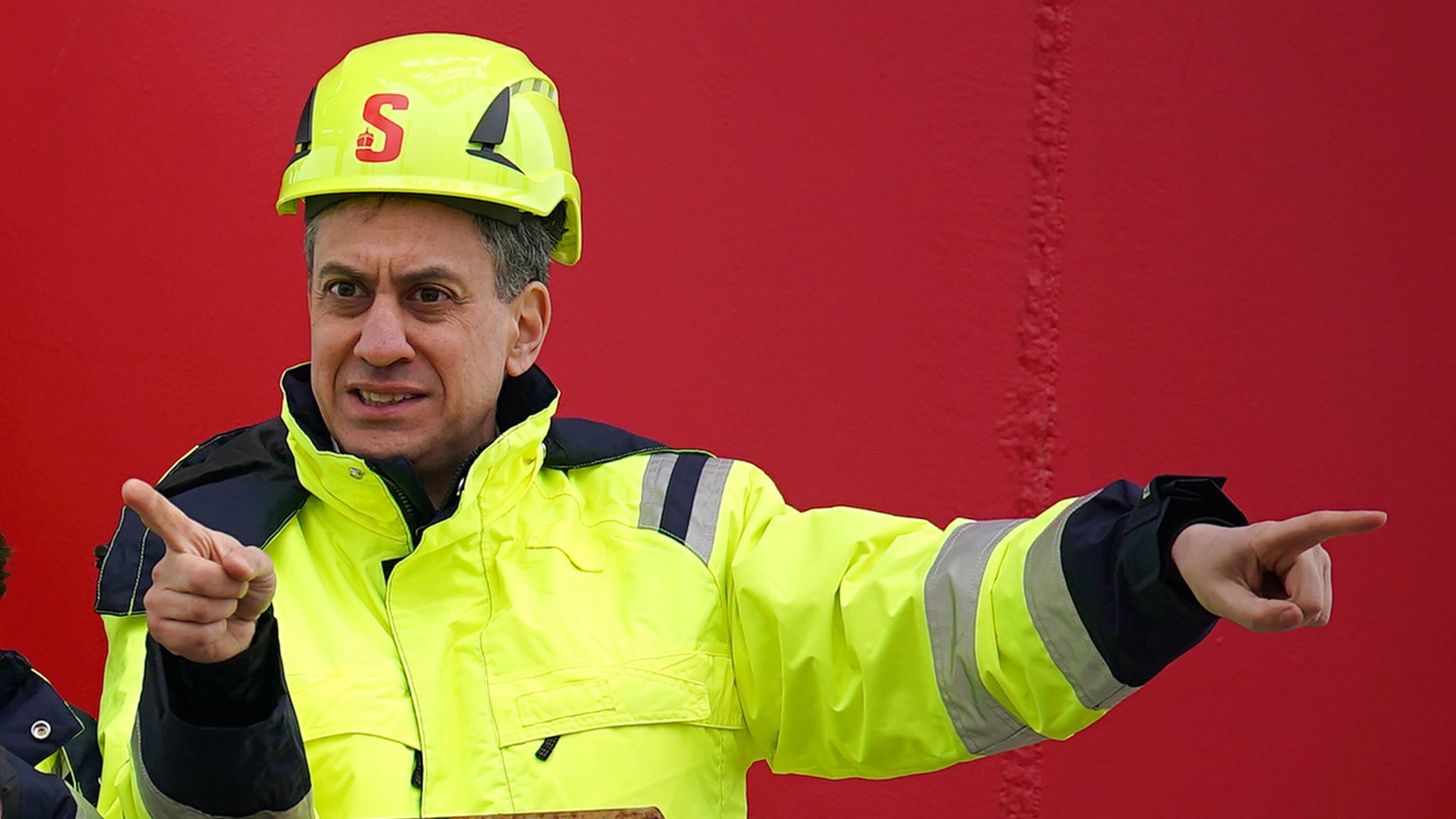 Ed Miliband wearing a hi-vis vest and helmet while speaking with construction workers on site.