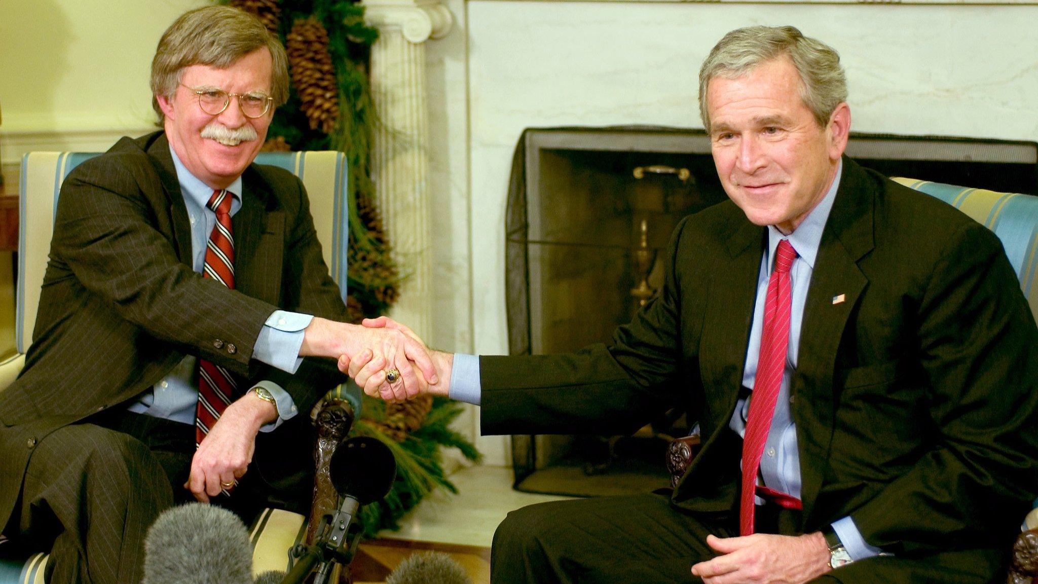 John Bolton shaking hands with President George W. Bush in the Oval Office, both smiling.