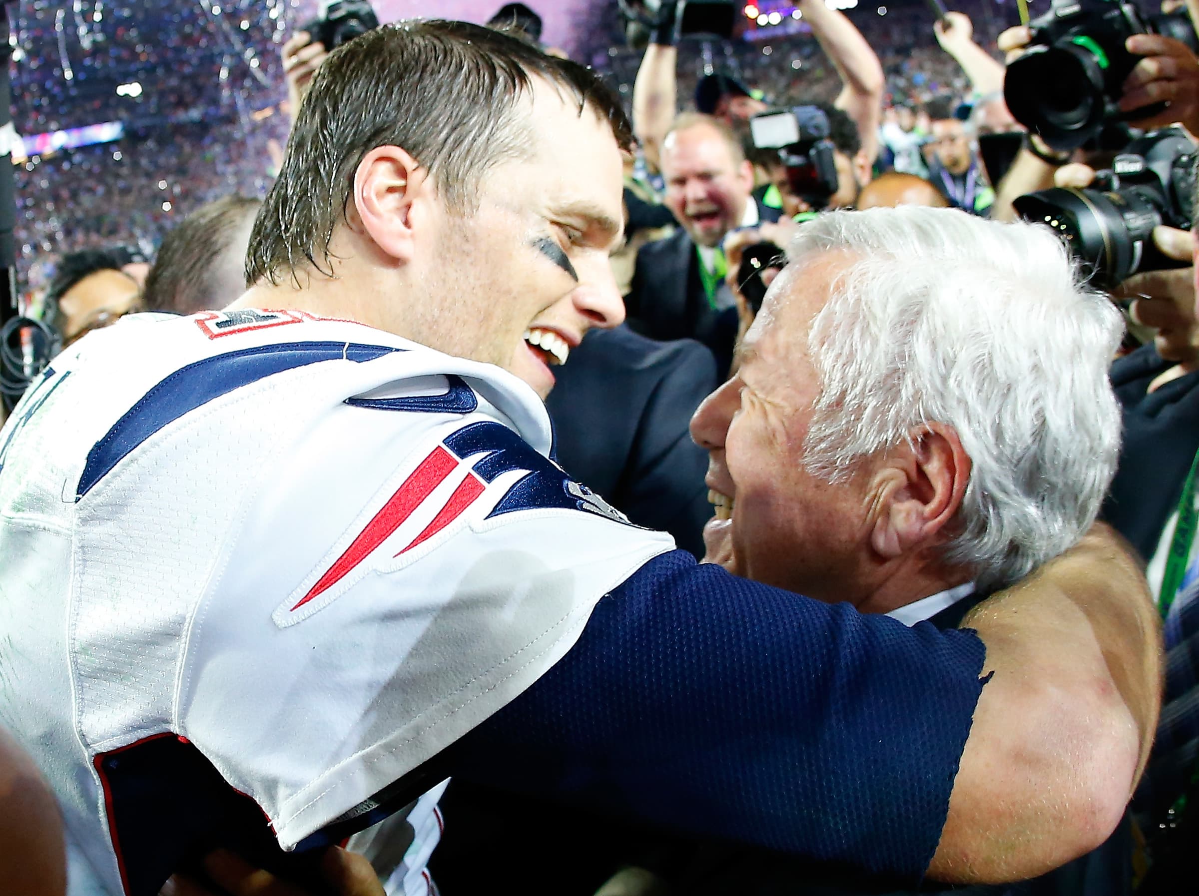 Robert Kraft hugging Tom Brady on the field, celebrating a Patriots victory with smiles and teammates around them.
