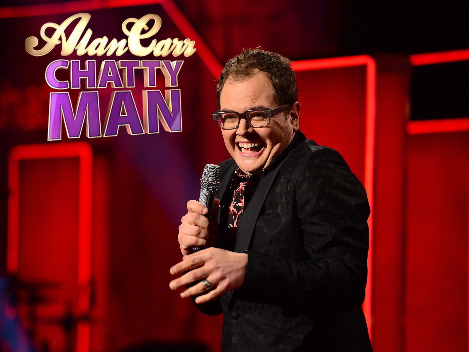 Alan Carr on the set of Chatty Man, engaging with the audience with a playful smile while seated at his iconic desk.