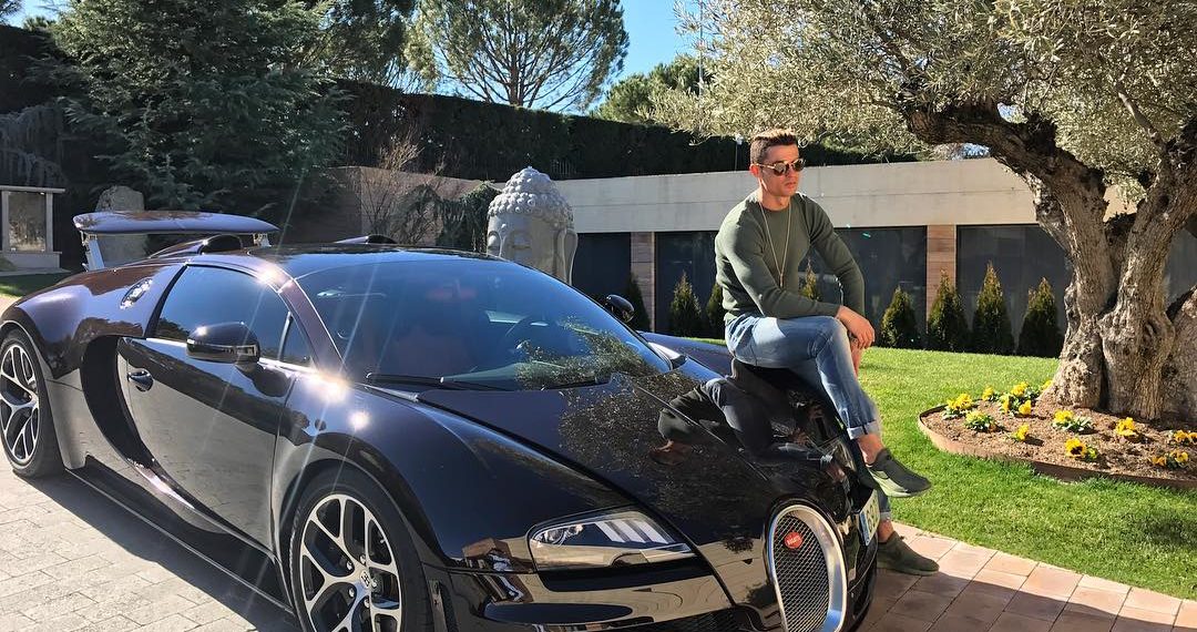 Cristiano Ronaldo sitting on the hood of his black Bugatti Veyron, posing confidently beside the luxury supercar.