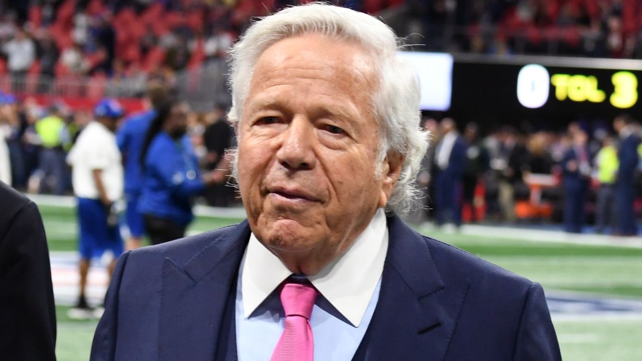 Robert Kraft in a suit being interviewed on the football field, speaking to reporters with stadium lights in the background.