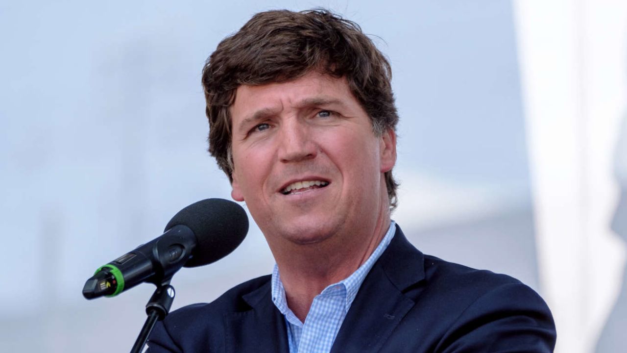 Tucker Carlson standing behind a microphone, poised to speak at a public event.