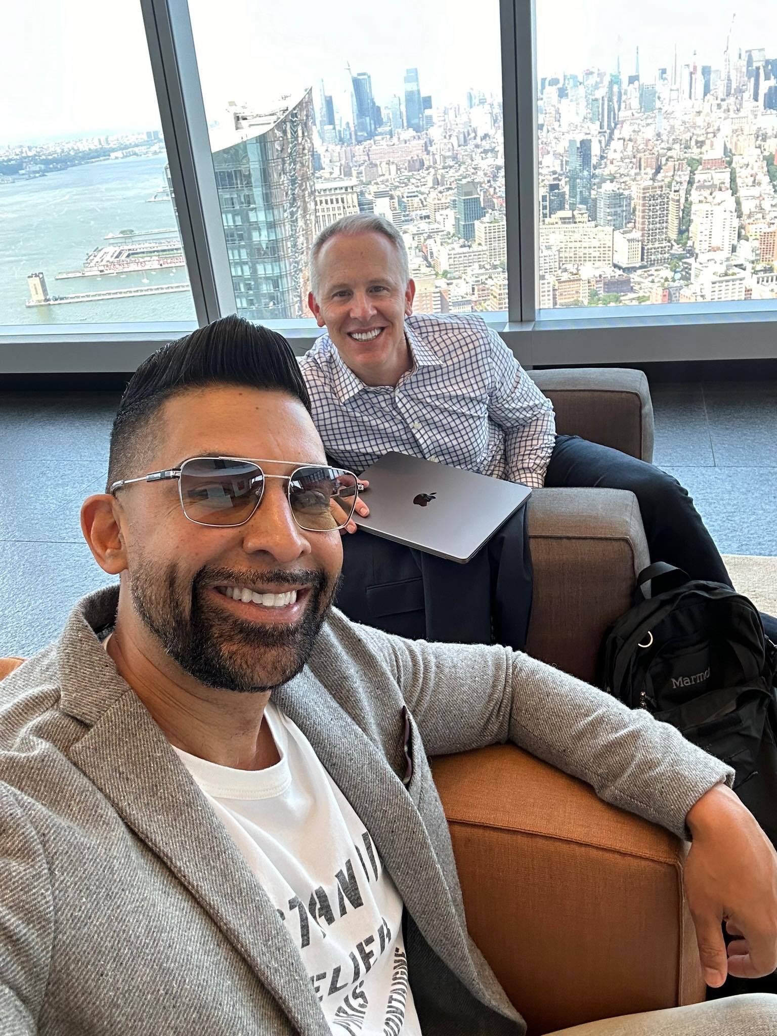 Dhar Mann and CEO Sean Atkins seated together, smiling in a modern penthouse office in New York, overlooking the city skyline.