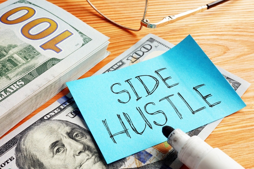 A blue Post-it note with the words “Side Hustle” resting on a stack of cash, symbolizing extra income opportunities.