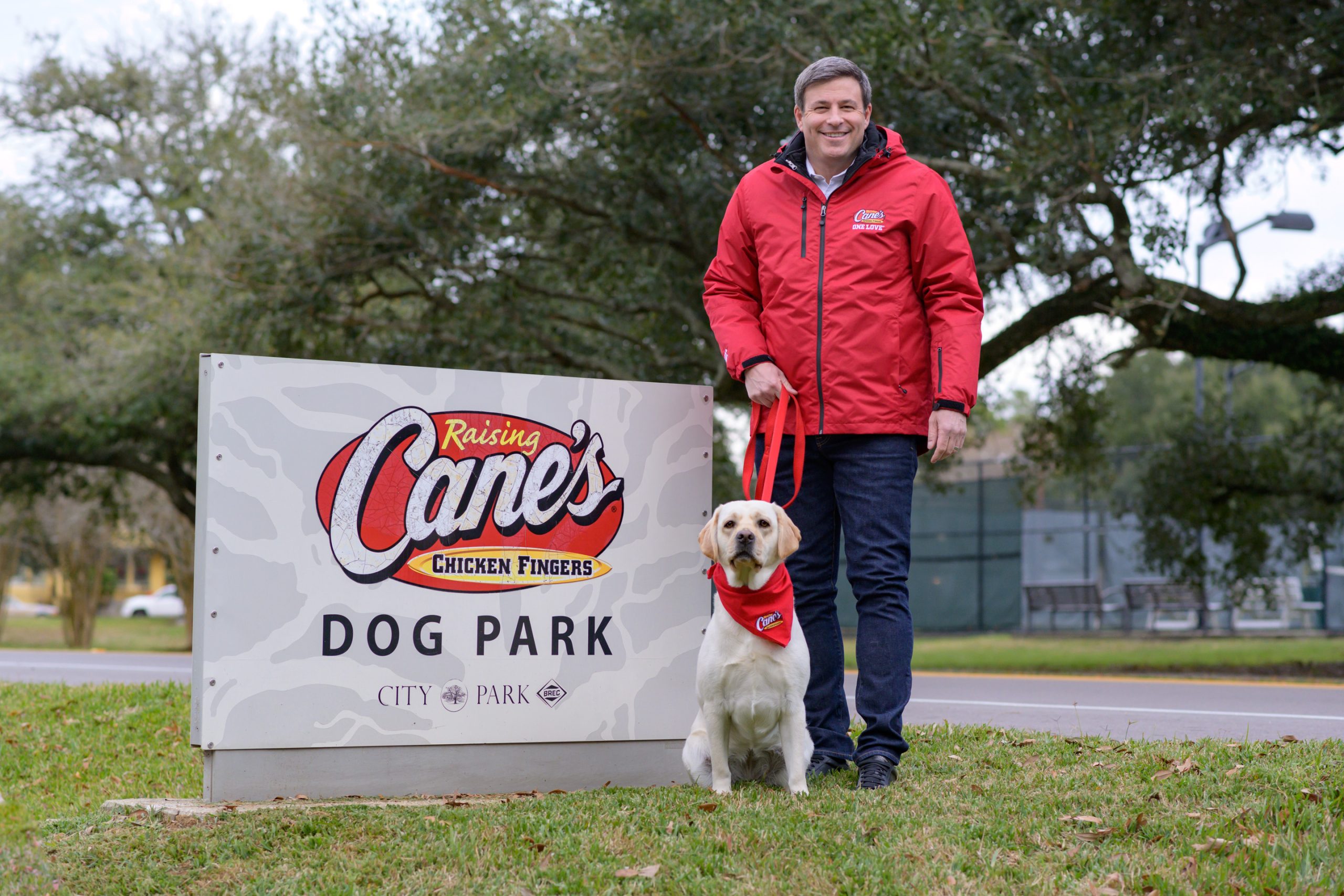 odd Graves standing beside a Raising Cane’s sign with his golden retriever, smiling proudly outside one of his restaurants.