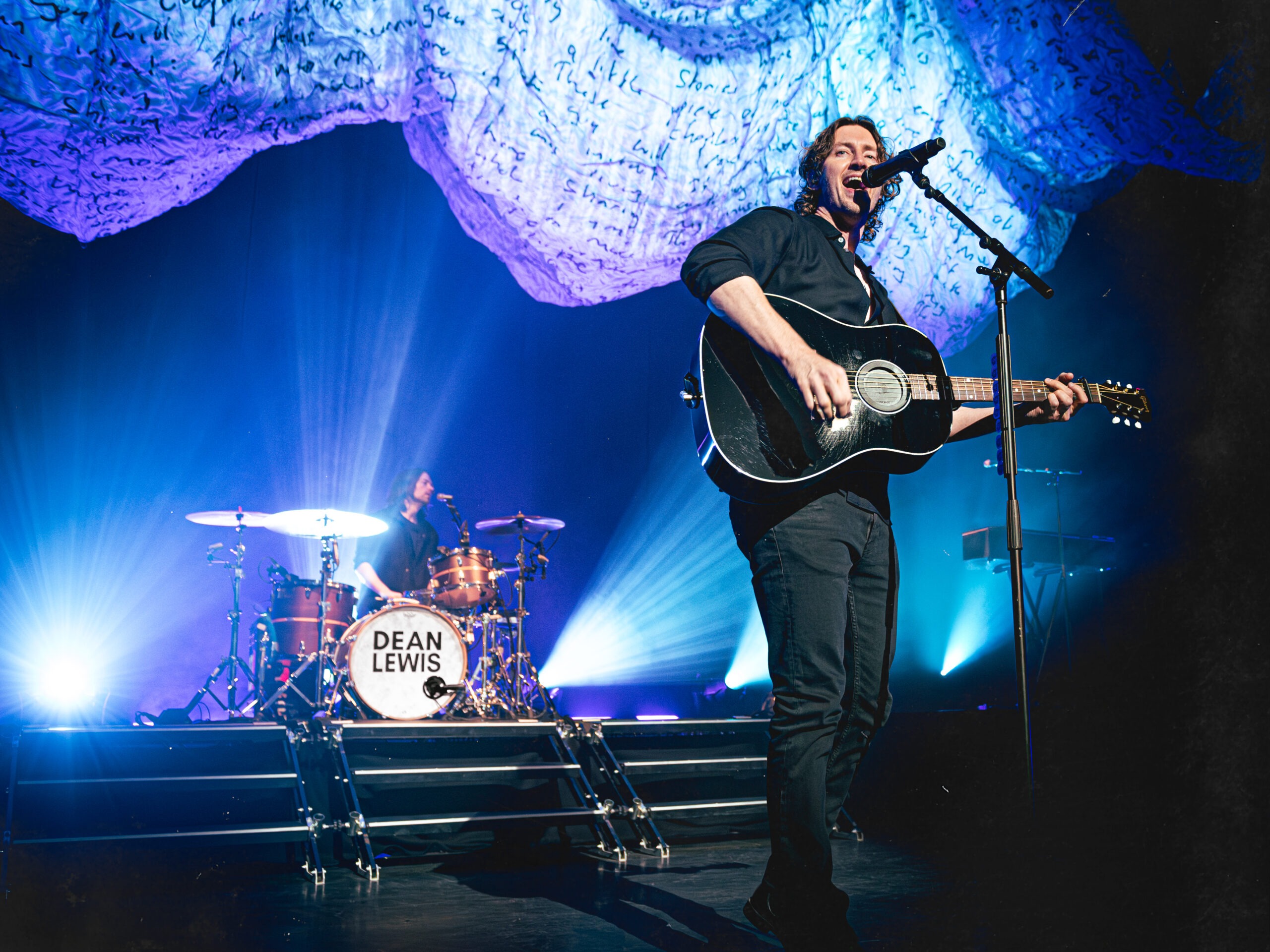 Dean Lewis performing on stage, singing into a microphone with stage lights illuminating him.
