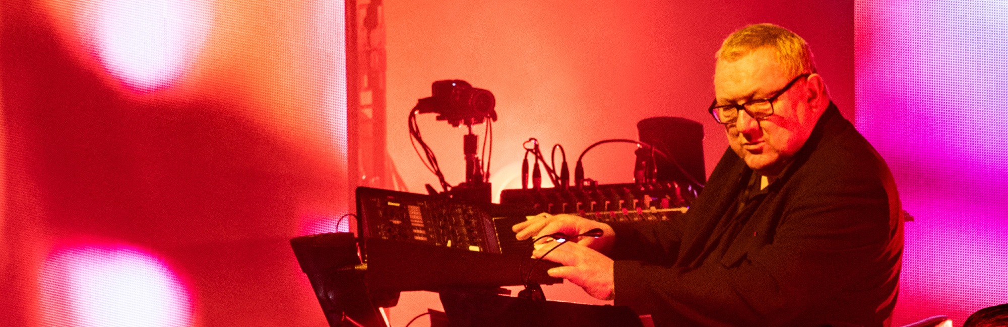 A photo of Dave Ball with his hands on the controls of a synthesiser or mixing desk, actively shaping sound and creating music.