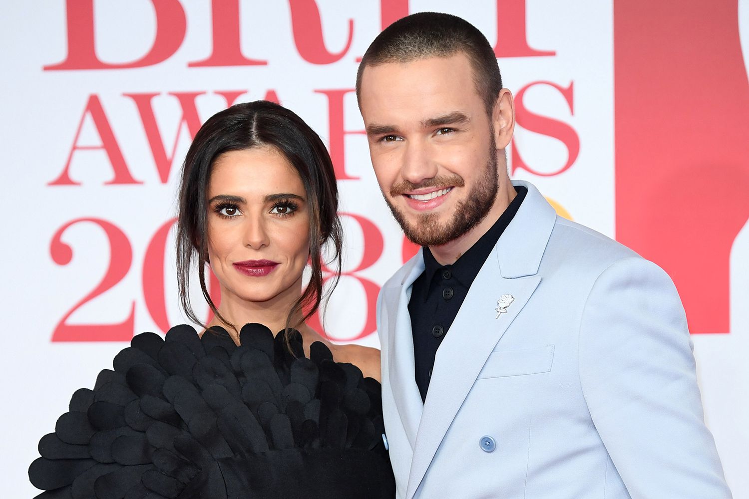 Liam Payne and Cheryl Cole posing elegantly on the red carpet, both in sleek black attire, smiling confidently with arms linked under bright spotlights and a glamorous backdrop of event signage.