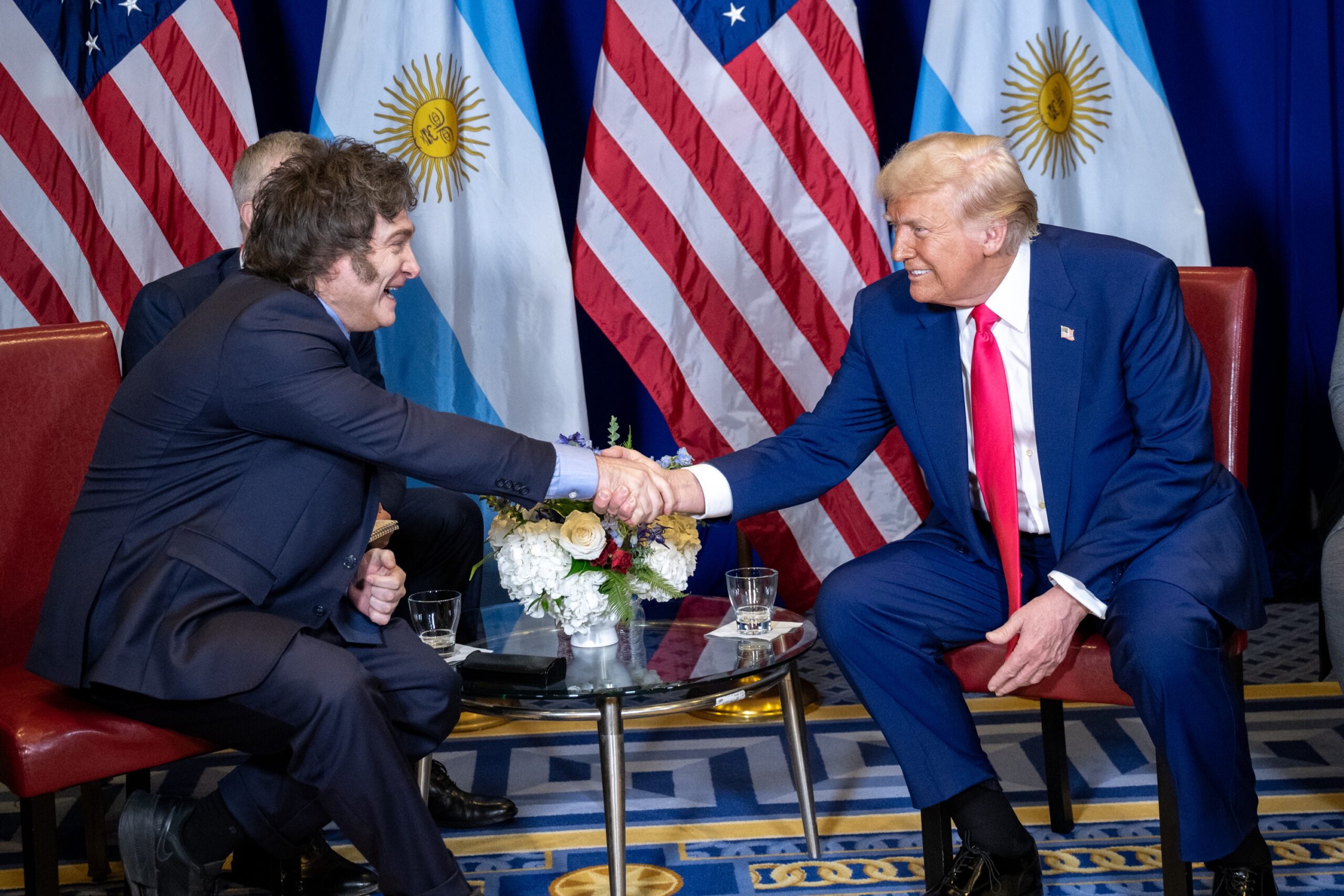 Argentine President Javier Milei and former U.S. President Donald Trump seated, shaking hands in front of the media during a White House meeting.