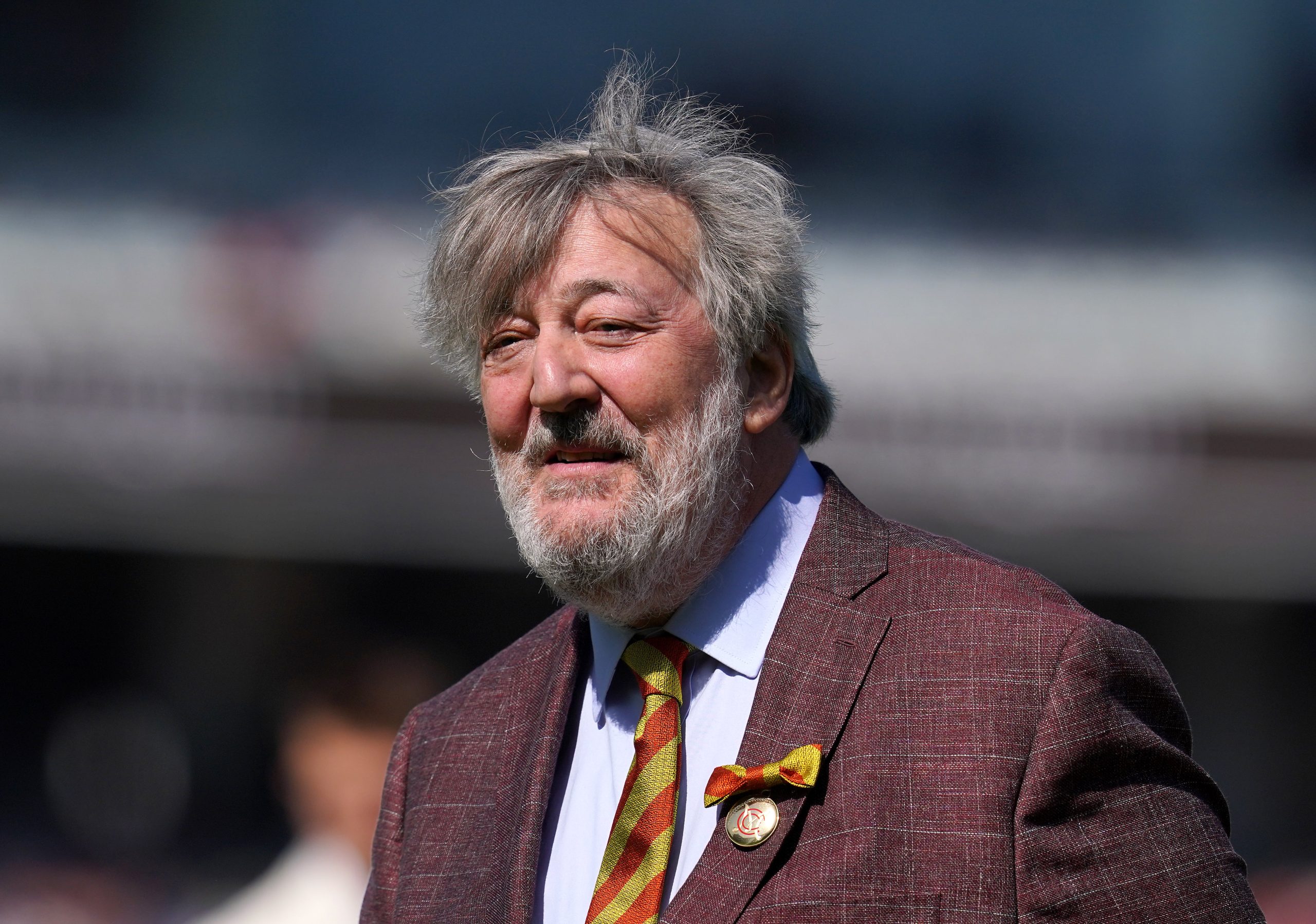 Stephen Fry standing outdoors wearing a maroon tweed blazer, smiling at the camera against a leafy background.