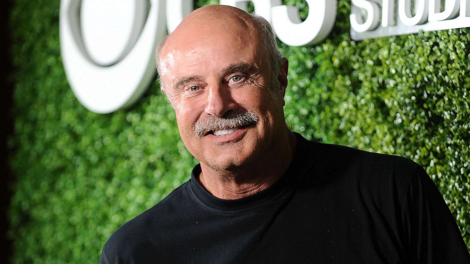 Dr. Phil McGraw smiling at the camera with a neatly trimmed green hedge in the background.