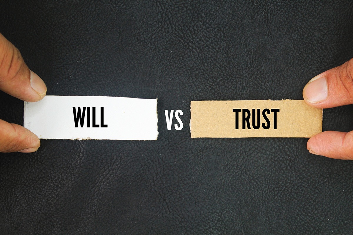 Two hands holding signs labeled “Will” and “Trust” with “vs” in between, symbolizing estate planning choices.