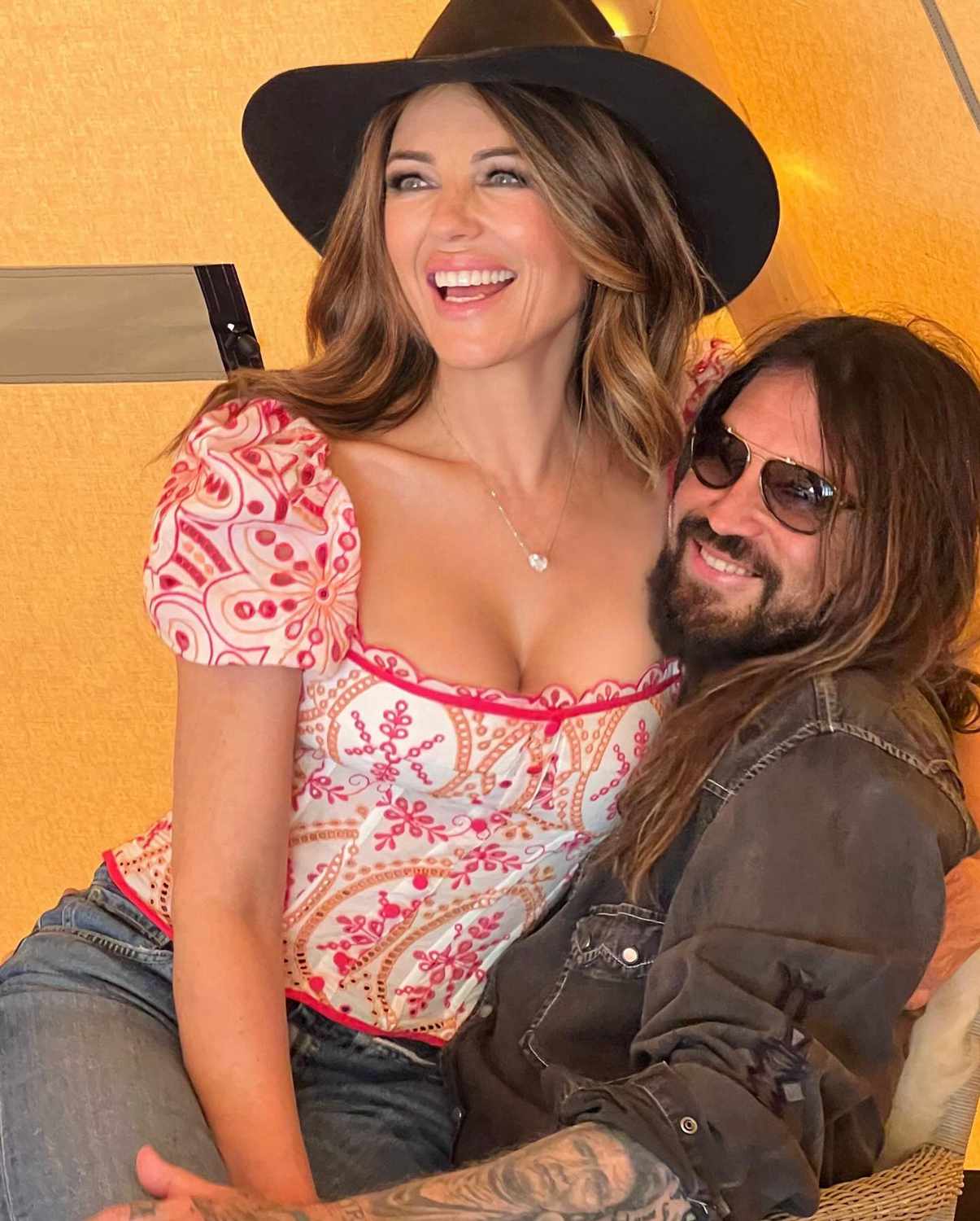 Elizabeth Hurley sitting on Billy Ray Cyrus’s lap, both laughing, with Hurley playfully wearing his hat, capturing a candid and affectionate moment between the couple.