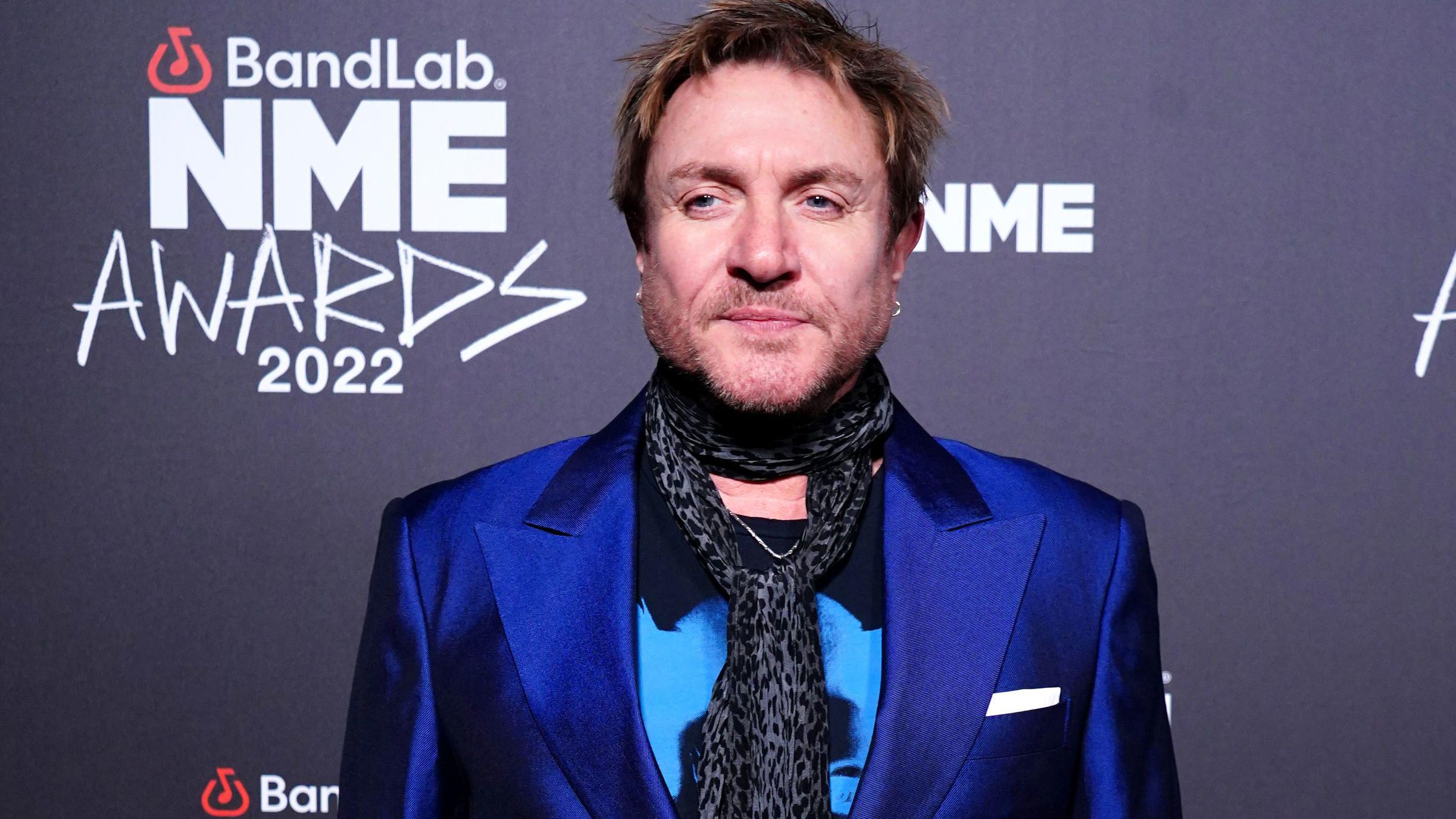Simon Le Bon attends the NME Awards 2022 wearing a blue satin blazer and patterned scarf, posing on the red carpet.