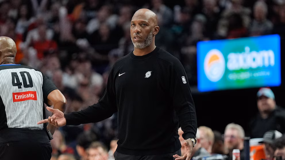 Portland Trail Blazers head coach Chauncey Billups reacts during an NBA game, standing courtside in a black team tracksuit as a referee walks past him.