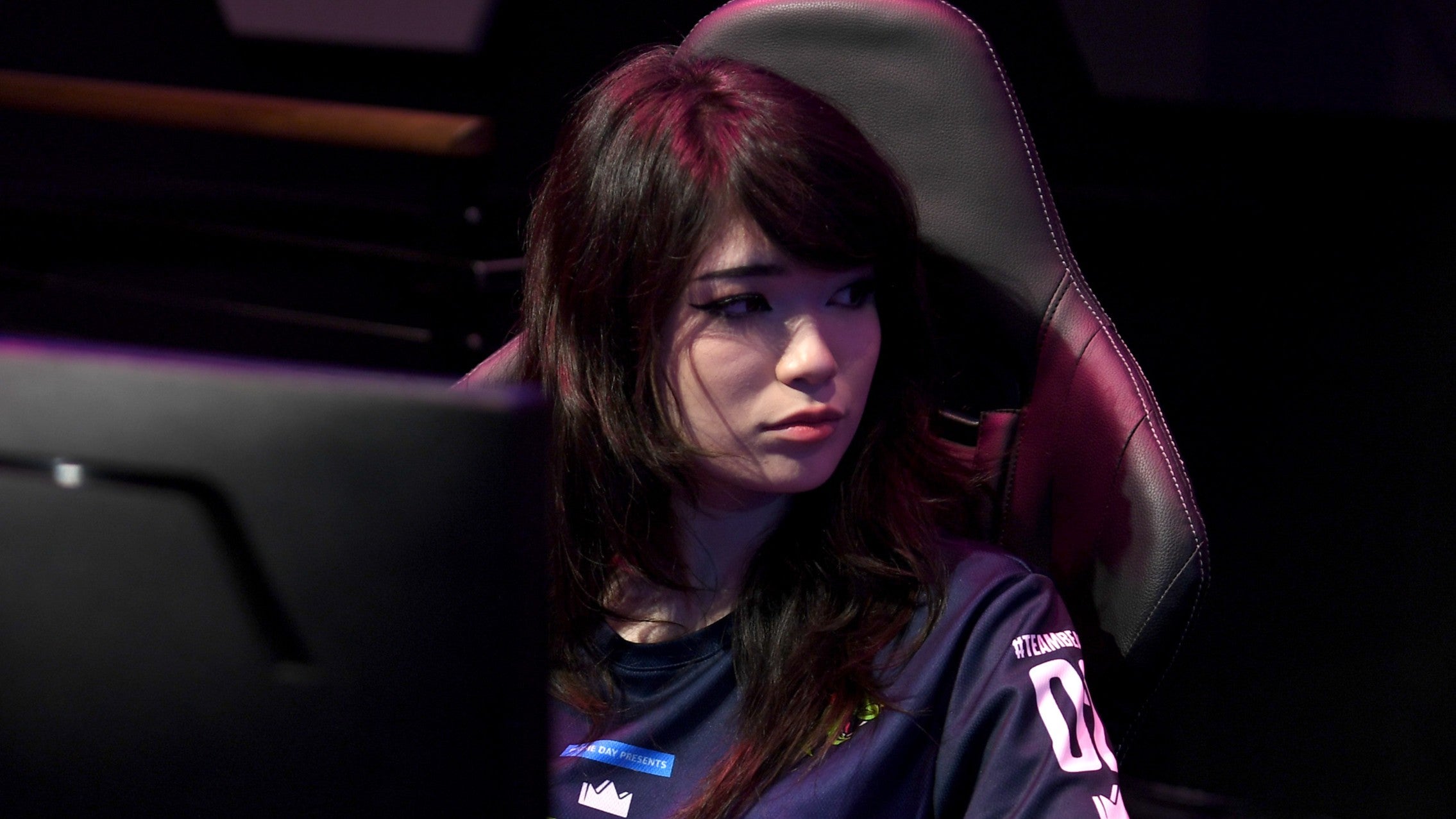 Emiru seated at her streaming setup during a live event, focused on her monitor with headphones on and lighting rigs illuminating her workspace.