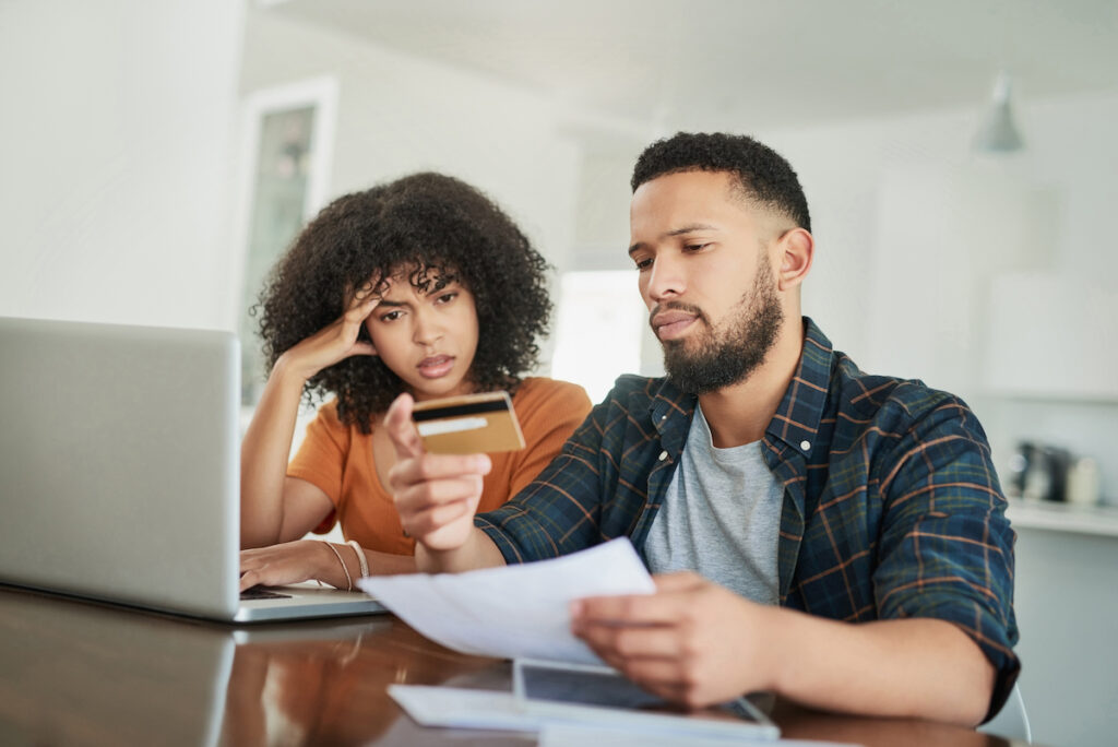 A couple sits at a table, holding a credit card and a financial document, looking confused and worried about their finances.