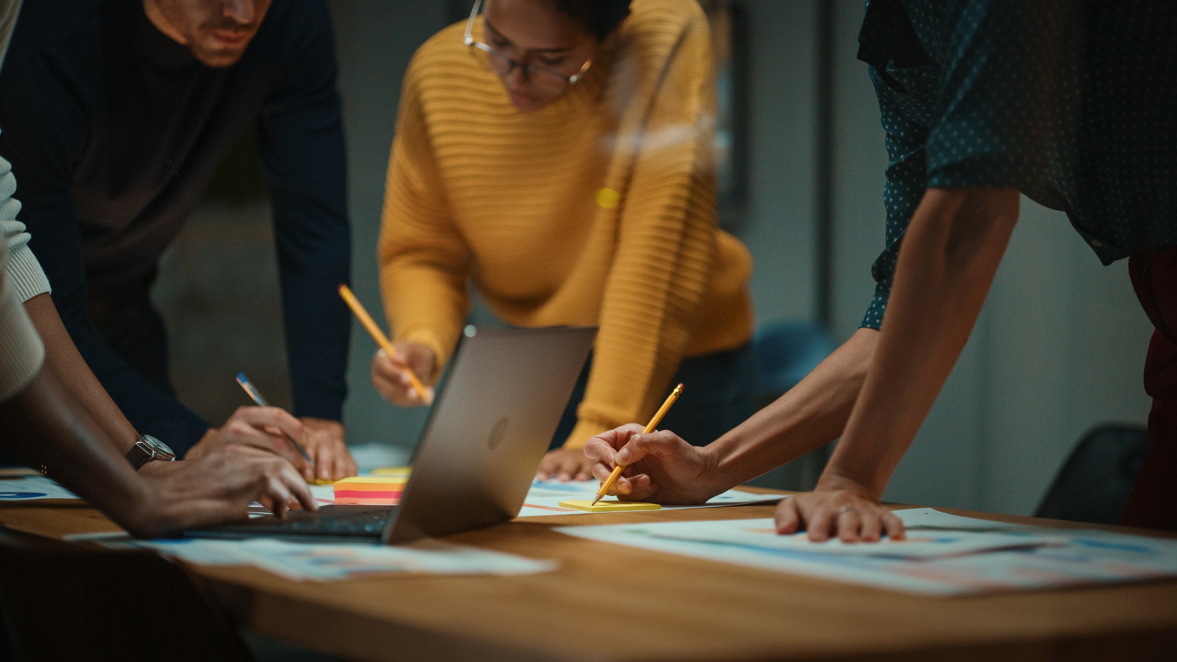 close up of diverse multiethnic team having conversation in meeting room in a creative office. colleagues lean on a conference table, look at laptop computer and make notes with pencils on notebooks.