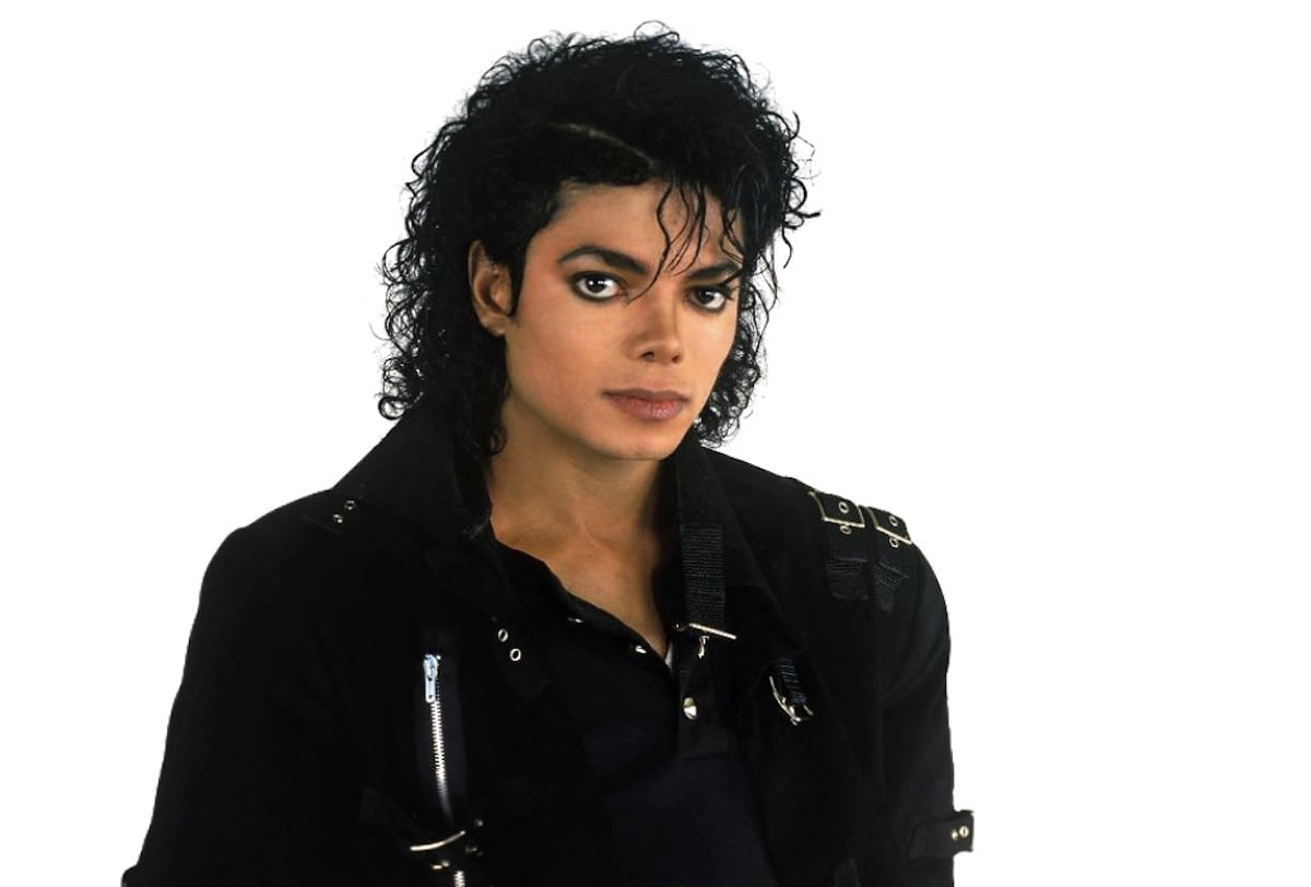 Michael Jackson wearing his iconic black leather jacket, standing against a white background, striking a classic pose.