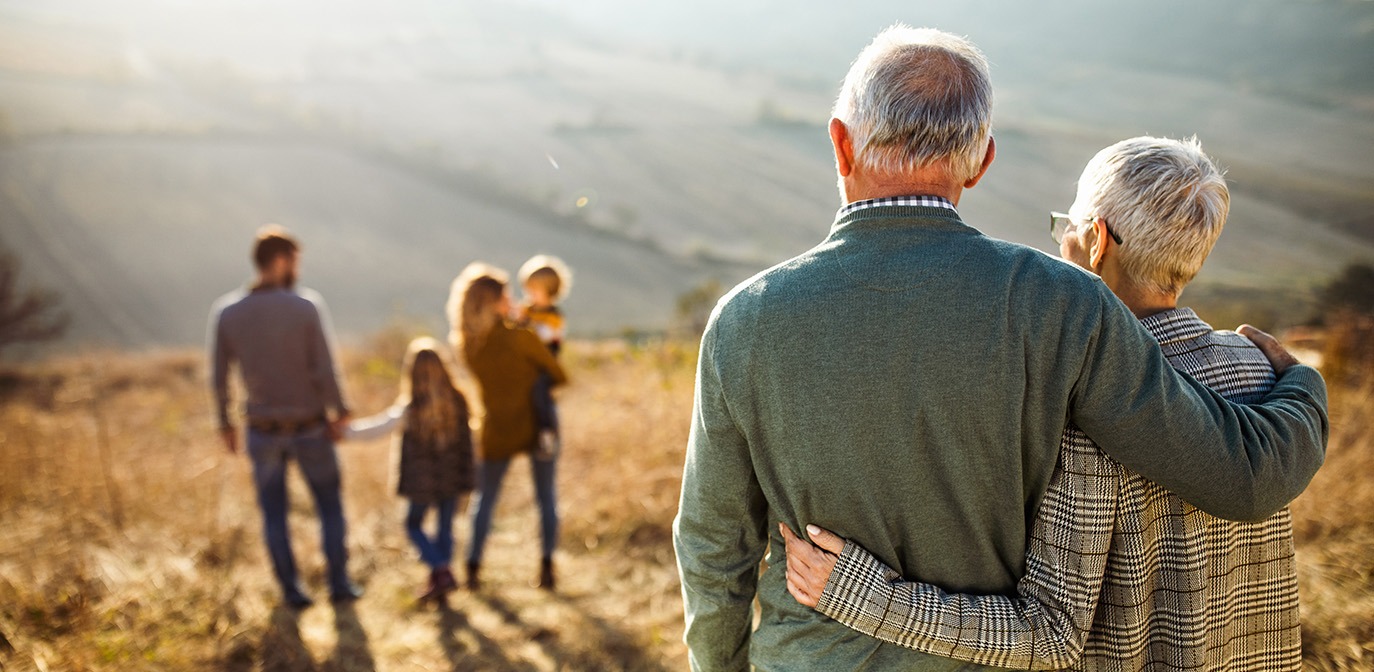 An older couple embracing while watching their children and grandchildren play together outdoors, symbolizing family legacy and generational wealth.
