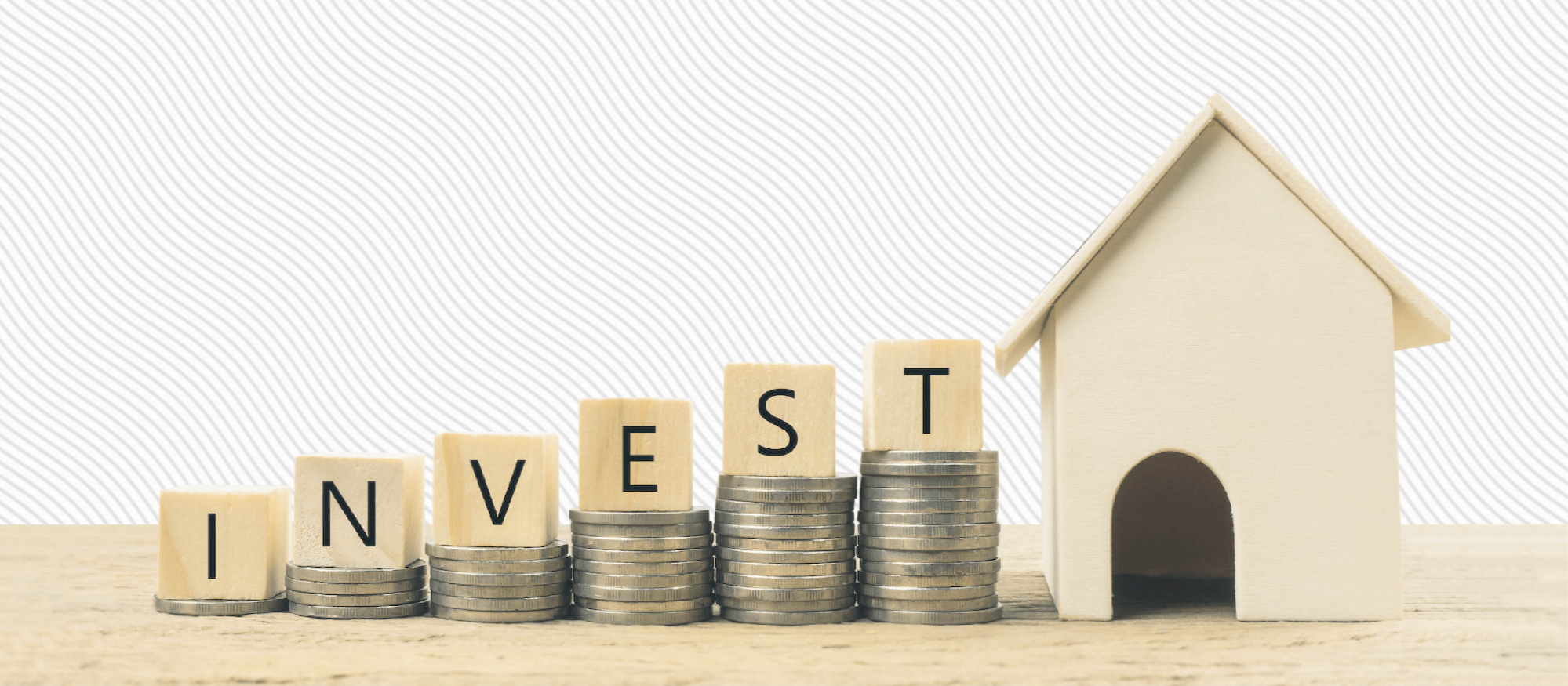 Wooden blocks spelling "INVEST" on stacks of coins next to a small wooden house, symbolizing real estate and financial growth.