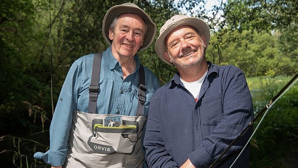 Bob Mortimer and Paul Whitehouse smiling while holding fishing rods beside a calm river.