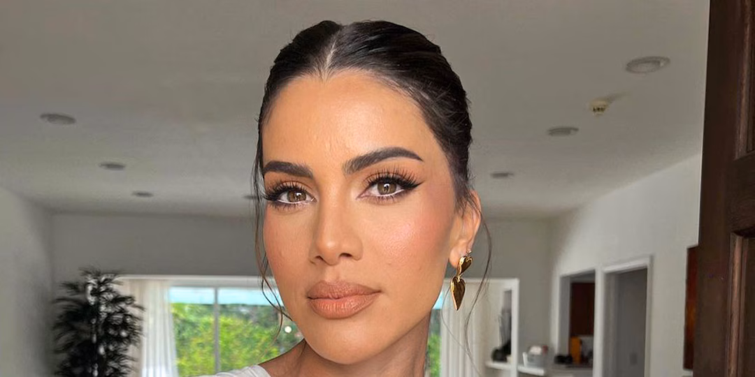 Close-up portrait of Brazilian fashion influencer and entrepreneur Camila Coelho, wearing natural glam makeup, gold heart-shaped earrings, and a white top, photographed indoors with soft daylight.