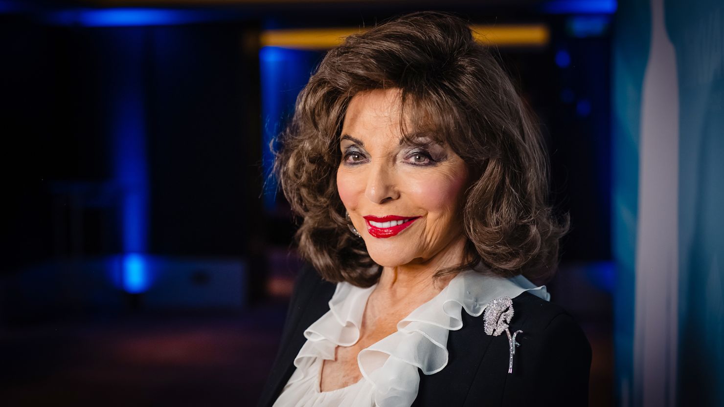 Dame Joan Collins smiling in an elegant black jacket with a white ruffled blouse, wearing a diamond brooch, photographed under soft blue lighting at a London event.