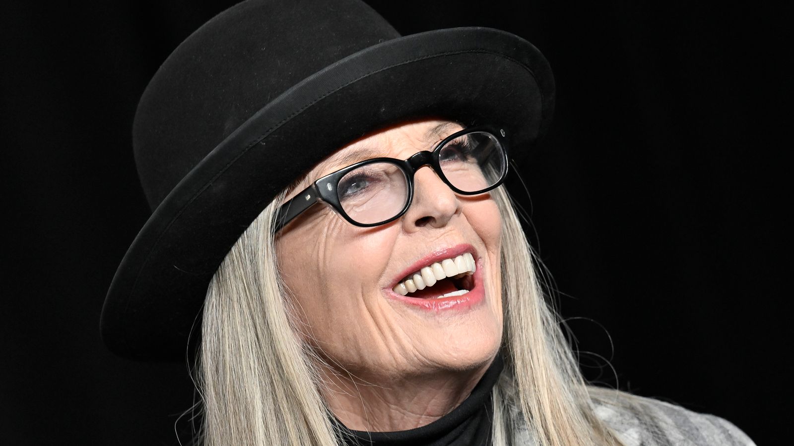 Diane Keaton smiling in signature black hat and glasses during a public appearance in Los Angeles.