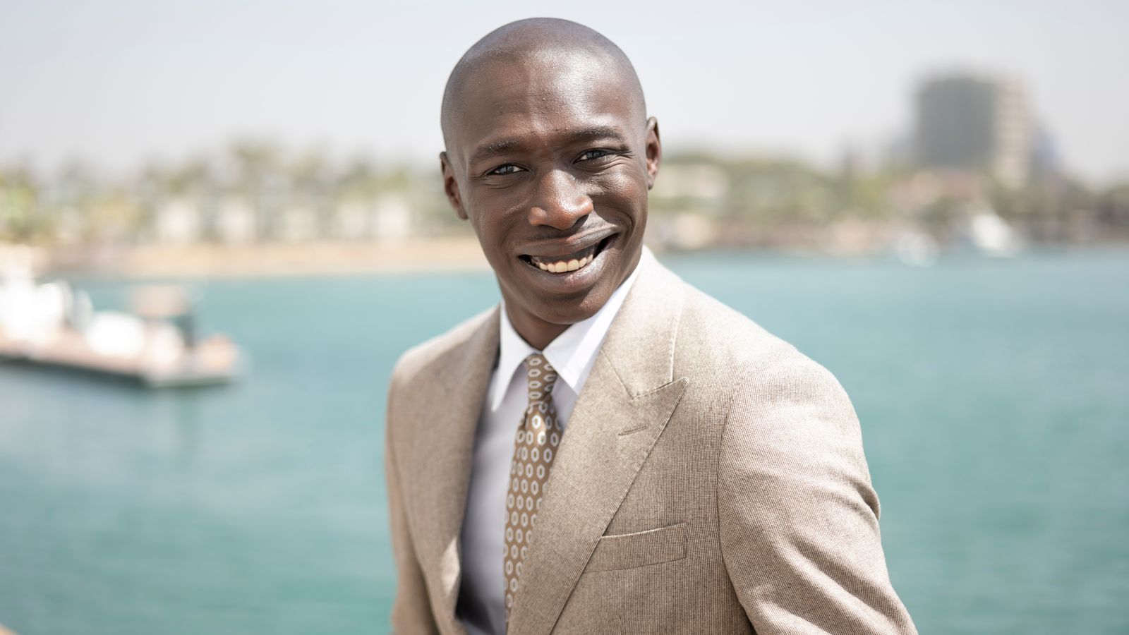 Khaby Lame standing by the sea, smiling casually with the ocean in the background.