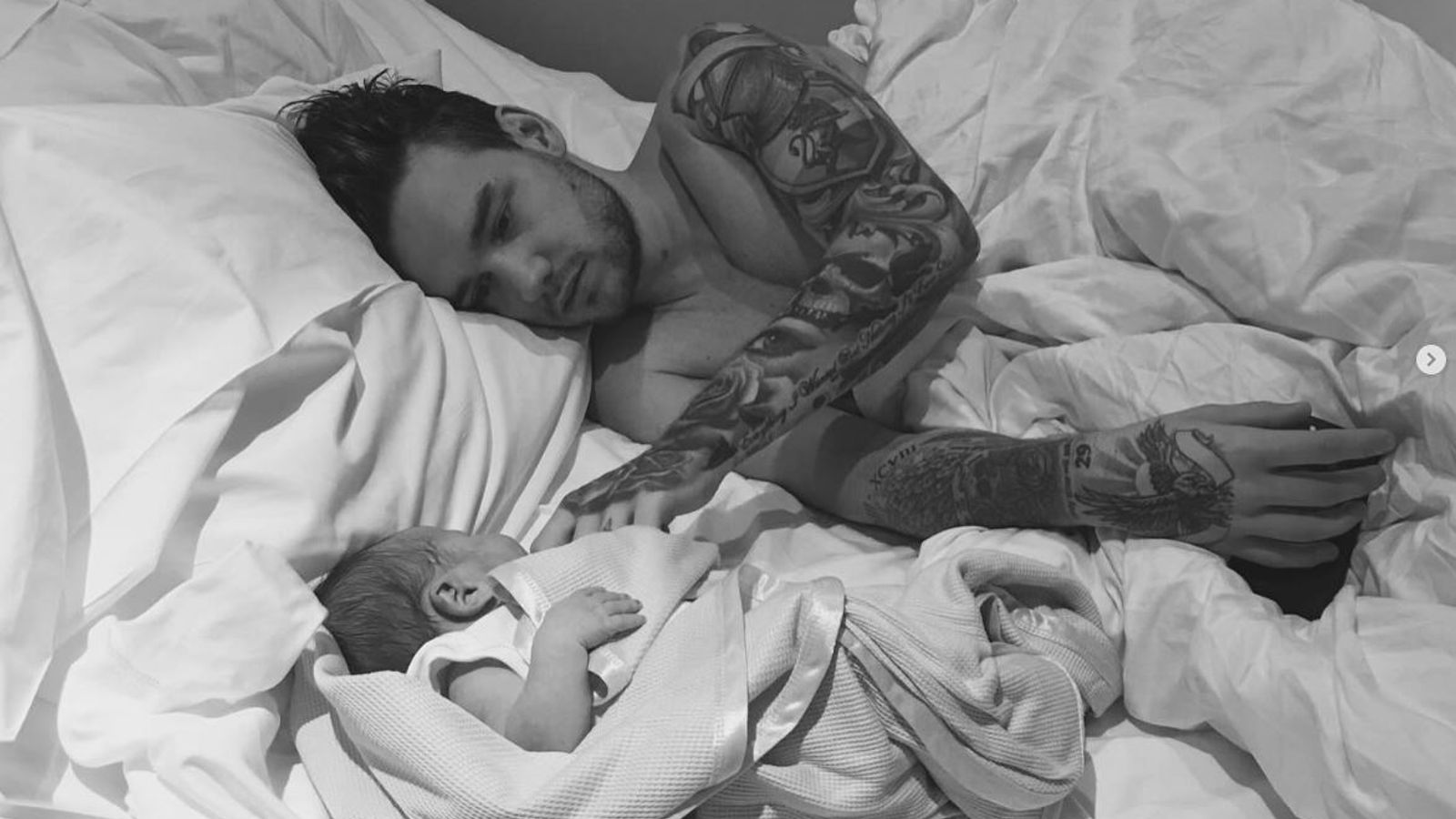Alt TextLiam Payne and his infant son Bear sharing a tender gaze in a cozy bed, with Liam cradling the baby gently against soft pillows and blankets, both illuminated by warm morning light filtering through sheer curtains.