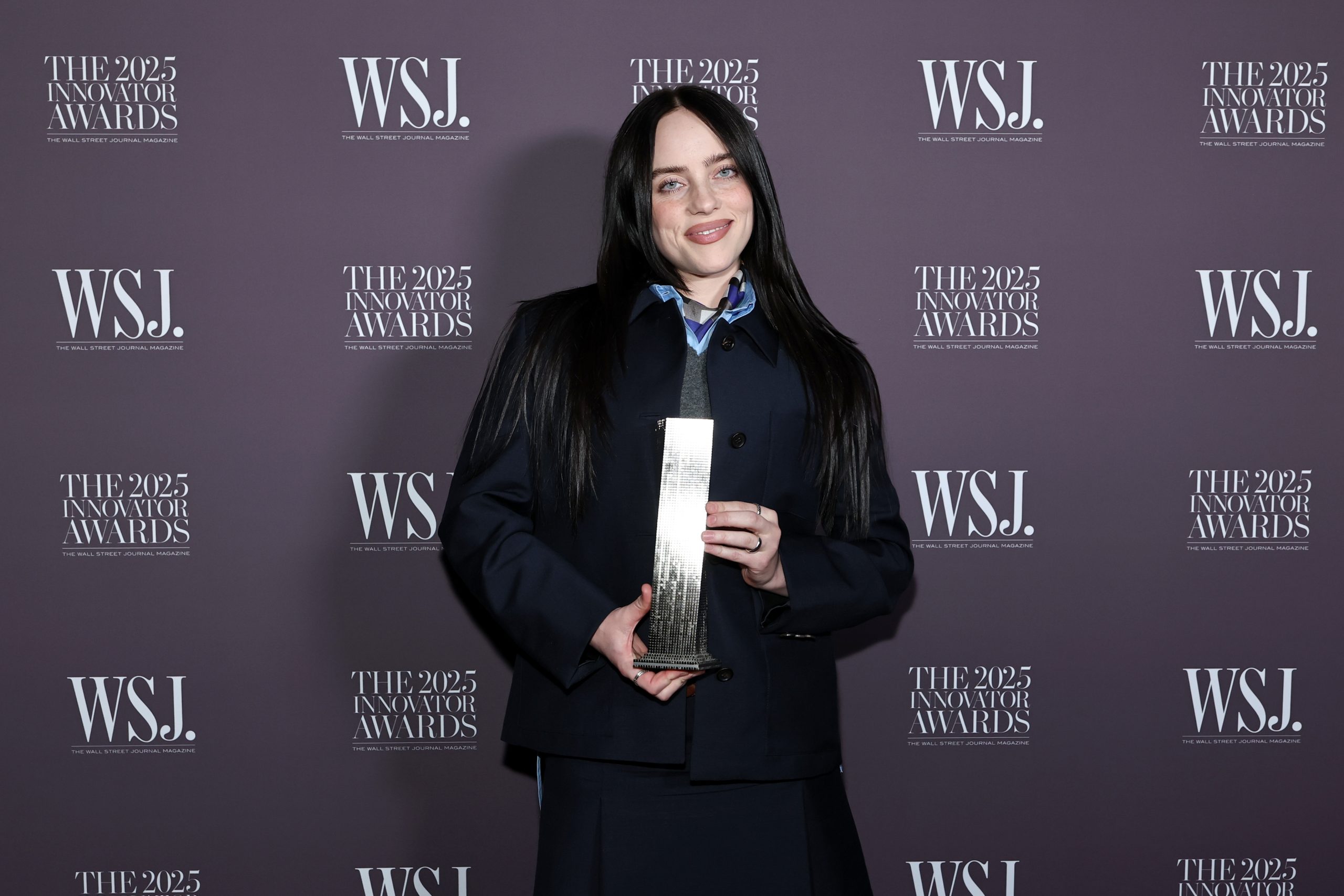 Billie Eilish holding her WSJ Innovator Award on the red carpet, smiling in a stylish outfit.